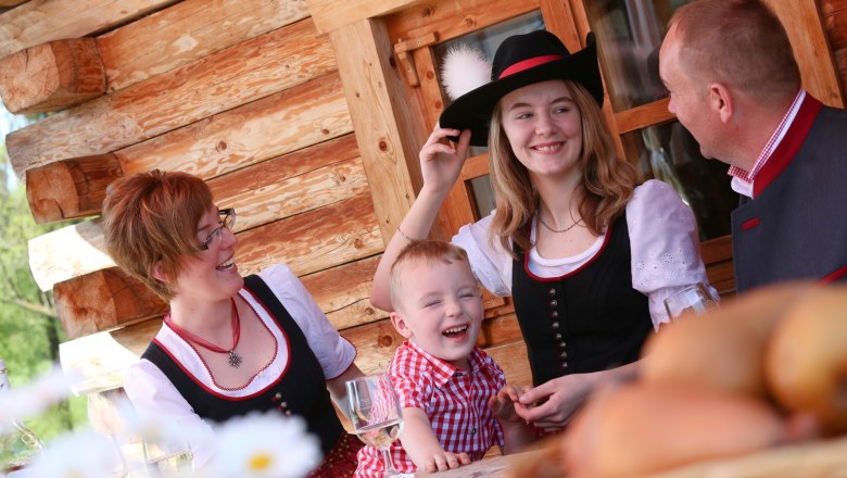Log cabin "D'Auszeit, © schwarz-koenig.at Family in traditional costume in front of a log cabin, laughing at the table.