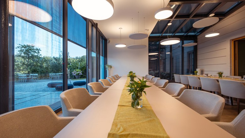 Refugium Hochstrass, © Niederösterreich Werbung / Maximilian Pawlikowsky Modern conference room with long tables, beige chairs and large windows overlooking a garden.