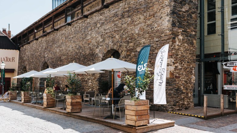 Café Wohnzimmer Gastgarten, © FMS Steinertor Gastronomie BetriebsgmbH Outdoor terrace of a café with parasols and plants in front of an old stone wall.