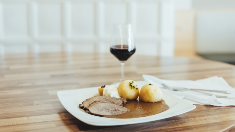 Always a favorite: Roast beef, © Niederösterreich Werbung/David Schreiber A plate of roast beef, dumplings and gravy on a wooden table, with a glass of red wine next to it.