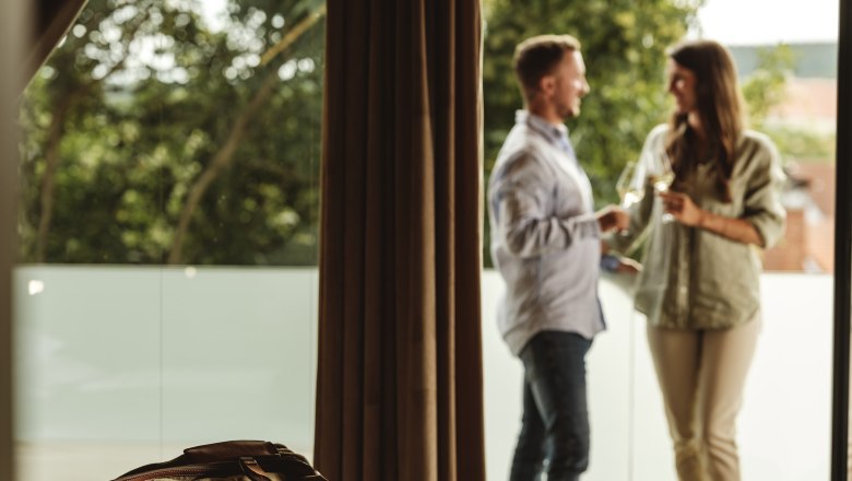 Winery Hagn Kellerstöckl, © Weinviertel Tourismus GmbH A couple is standing on a balcony holding glasses of wine, a travel bag lies on a bed in the foreground.