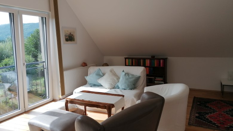 Living area in the "garden room", © Wiener Alpen Bright living area with sofa, armchairs, bookshelf and large window with a view of the greenery.