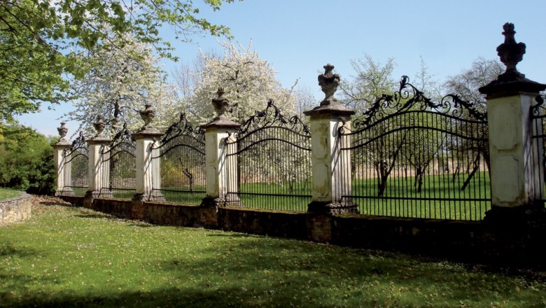 rg_fence_cutout_c_lock_greillenstein_gary_rogers, © Renaissanceschloss Greillenstein/Gary Rogers A decorative iron fence with stone pillars and ornate spikes in front of flowering trees in the background.