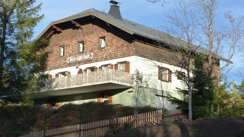 The refuge, © Mario Putz A traditional building with wooden cladding and balcony, surrounded by trees and a wooden fence. Sign: "Döhler shelter".