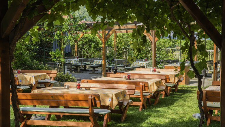 Heurigen garden, © Monika Loeff A wine tavern garden with wooden benches and tables under an arbor of vines.