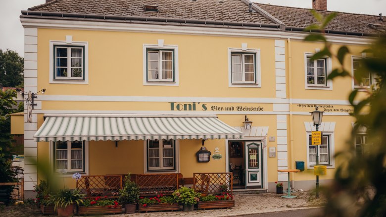 Inn with history in Kilb, © Niederösterreich Werbung/Daniela Führer Yellow building with awning and plants, labeled 'Toni's Bier- und Weinstube'.