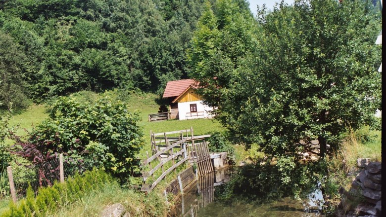 Pichlmühle, © Gemeinde Loich A small hut with a red roof stands in a green landscape next to a stream and trees.