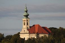 Parish church of St. Nicholas, Wolfpassing, © Gemeinde Hochleithen Parish church of St. Nicholas in Wolfpassing with red roof and green tower in front of a cloudy sky.