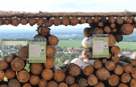 Balcony Tullnerfeld, © Gemeinde Tulbing, Thomas Gattinger View through stacked logs onto a landscape with fields and houses.