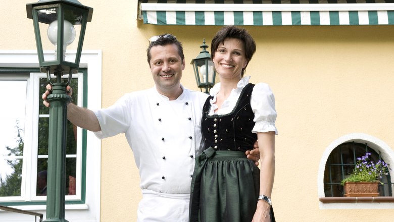Gasthof Mitter, © Christa Flach A man in chef's clothes and a woman in traditional costume stand smiling in front of a yellow building with green lanterns.