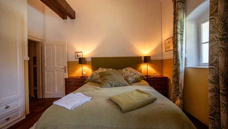 Forester's lodge Langschlägerwald, © Niederösterreich Werbung / Maximilian Pawlikowsky Cozy bedroom with double bed, green bed linen and wooden decor. Two bedside tables with lamps, window with curtains, wooden beams on the ceiling.