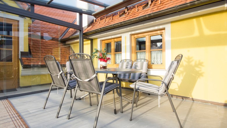Cozy terrace, © Winzerhof Semmler Winter garden with table and chairs, yellow walls, glass roof.
