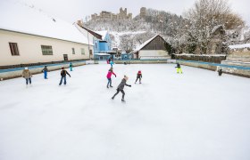 Kirchschlag ice rink, © Wiener Alpen, Martin Fülöp People skating on a square in Kirchschlag, with a castle ruin on a hill in the background.
