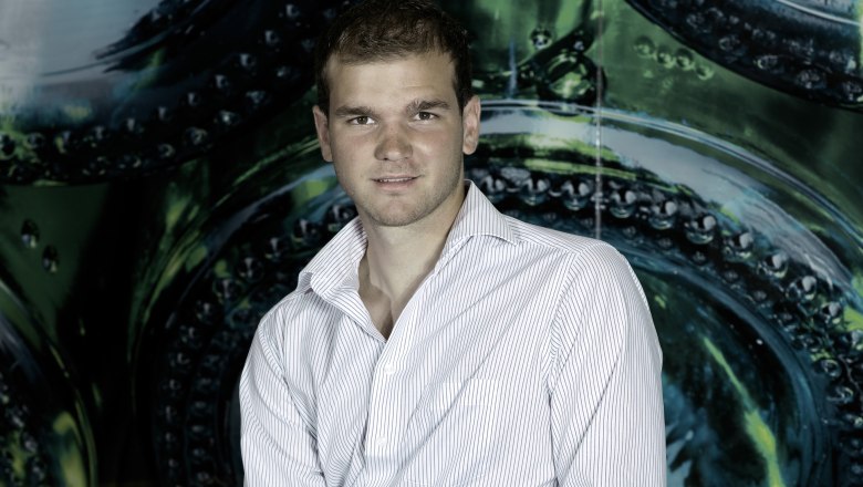 Winemaker Martin Kohl, © Weingut Kohl A man is sitting on a table labeled 'Weingut Kohl', holding a wine glass and a wine bottle next to him.