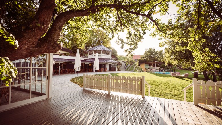 Distant view, © Matthias Kronfuss A garden with a terrace, trees and a pavilion in the sunlight.