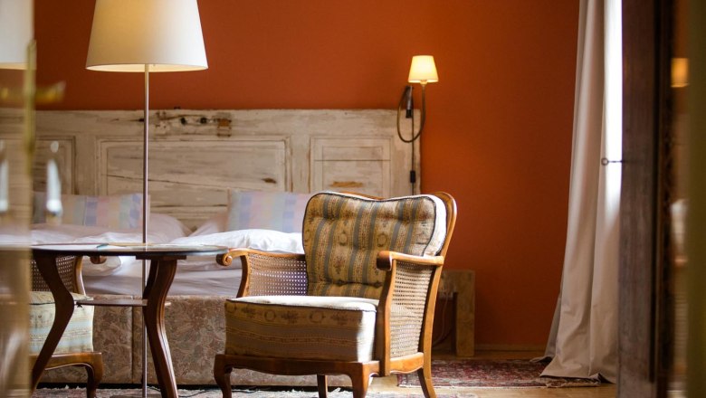 Klinger's guest house, © Astrid Bartl Cozy room with armchair, table and floor lamp in front of a bed with rustic wood paneling.