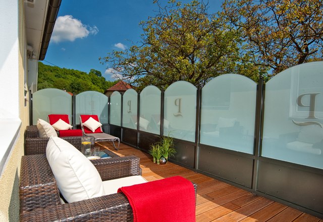 Wellness terrace, © Residenz Wachau A terrace with rattan furniture, red blankets and cushions, surrounded by glass walls and trees in the background.