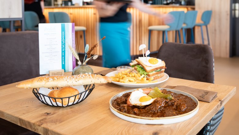 Regional and seasonal dishes, © Alexander Künzl Photography A table in a restaurant with regional dishes: a plate of goulash and fried egg, a sandwich with chips and a bread basket.