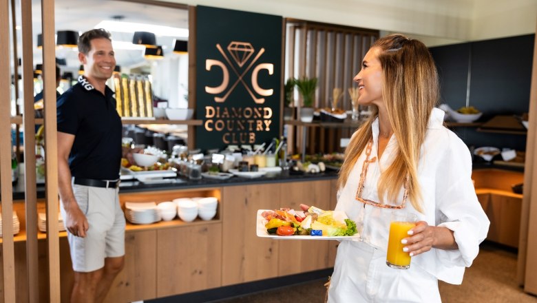 Hotel DCC, © Diamond Country Club Man and woman standing in front of the breakfast buffet. The woman is holding an orange juice and a plate of food in her hands.
