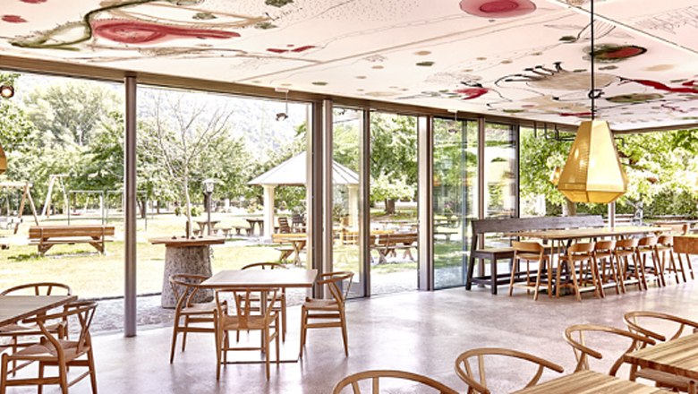 Interior view, © Familie Mang Interior view of a modern café with large windows and wooden furniture, overlooking a green outdoor area.