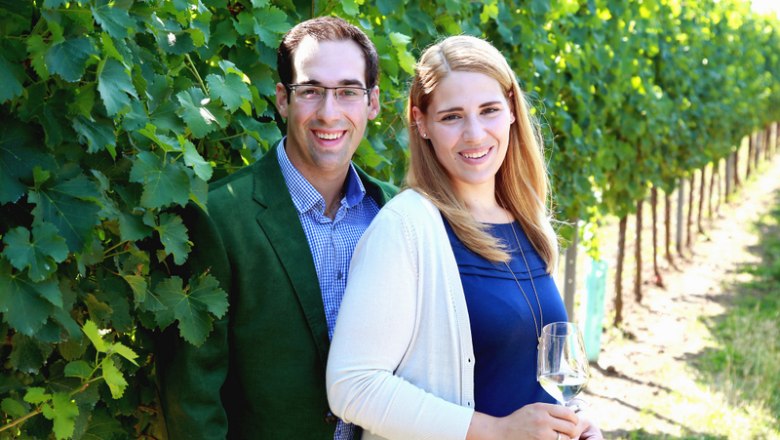 Winegrower couple in the vineyard, © Lavinia A smiling couple stands in a vineyard, the woman holding a glass of wine.