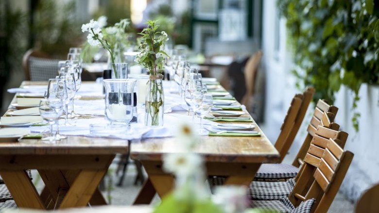 Gebeshuber, © Gebeshuber Long wooden table outside, set with glasses, cutlery and flowers.