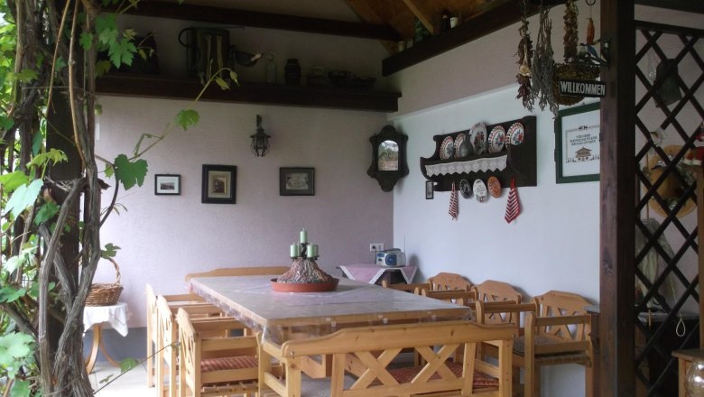 Guesthouse Schlossblick, Laube, © Marschik Covered terrace with table and chairs, in the background a wall with various wall decorations.