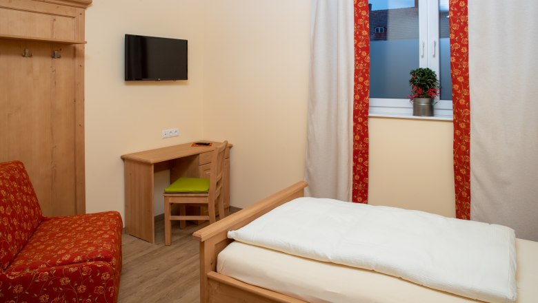 Single room, © Nadine Beles A cozy single room with wooden furniture, a bed, a desk and a TV on the wall. A window with curtains and a plant.