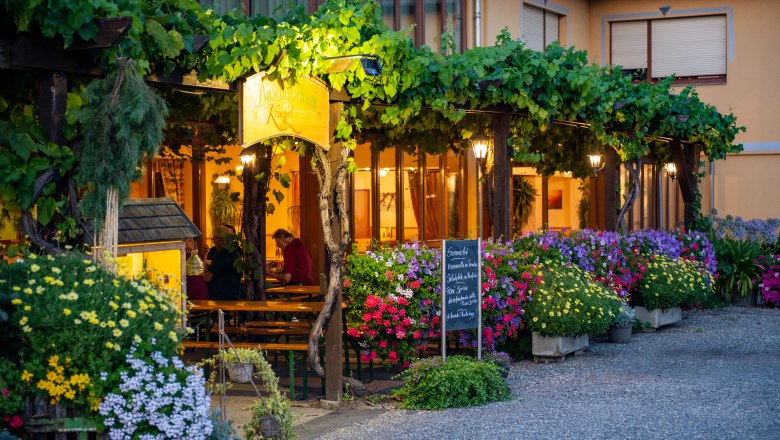 Guest garden, © Martin Lifka Photography A cozy garden with flowering plants and vines, illuminated by warm light. People sit at wooden tables under a pergola.