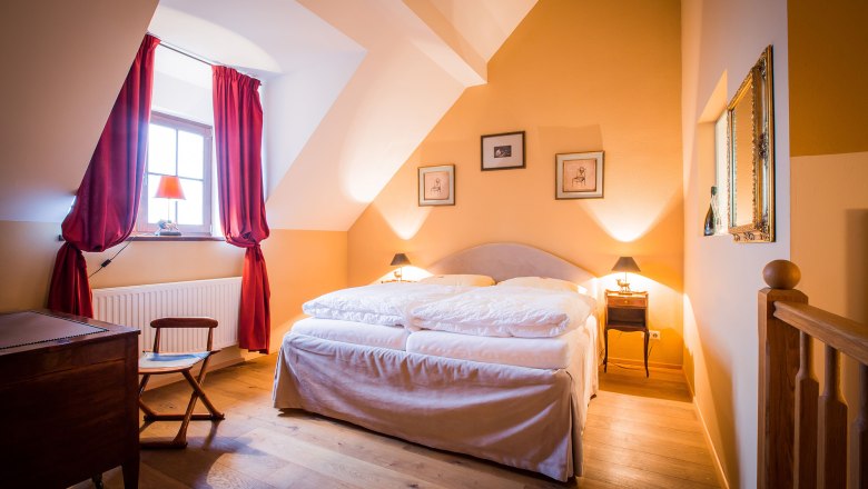 Deluxe double room with castle view, © Schlossschenke A cosy double room with a large bed, red curtains and warm colors. Pictures hang on the wall and a window lets in natural light.