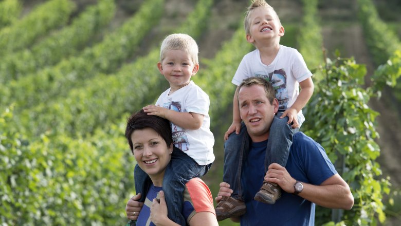 Holzmann family, © Weingut Holzmann Family in a vineyard, two children on their parents' shoulders.