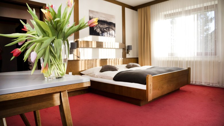 Double room, © Doris Seebacher A cozy double room with a wooden bed, red carpet and a table with a bouquet of tulips.