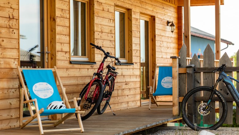 Sunny courtyard, © Wiener Alpen / Martin Fülöp Wooden veranda with two bicycles and blue deckchairs in front of a wooden house.