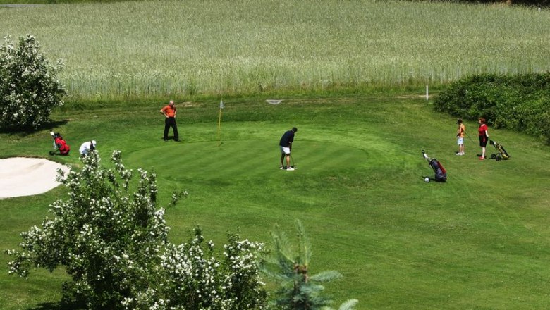 GC Herrensee, © GC Herrensee People play golf on a green course with trees and a sand bunker.