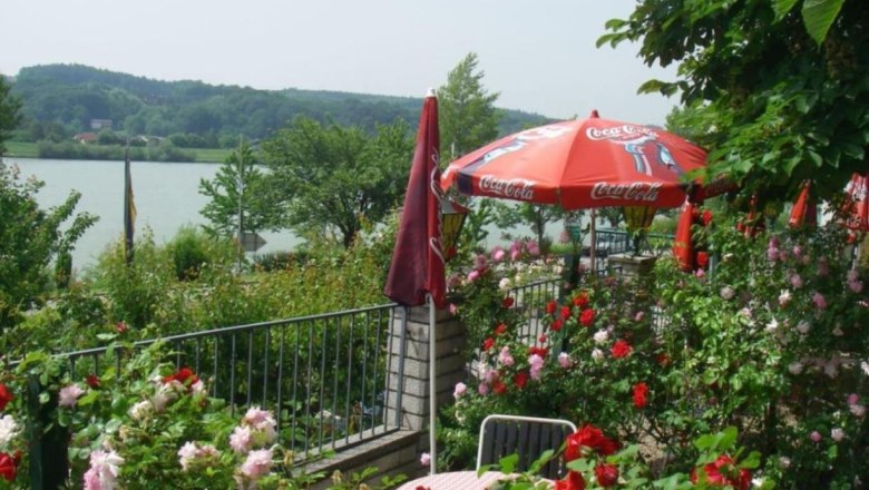 Garden, © Wachauerhof A blooming garden with red and pink roses, a table with chairs and a red parasol with the Coca-Cola logo, with a lake in the background.