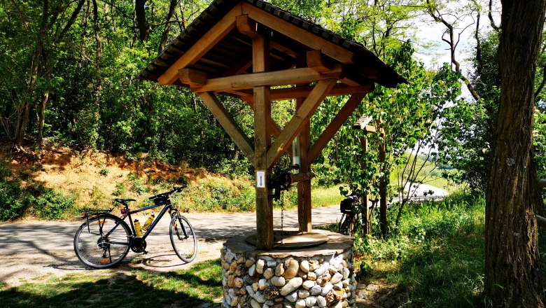 Wine fountain in Großwetzdorf, © Weinstraße Weinviertel A wooden wine fountain with a bicycle next to it, surrounded by trees and green foliage.