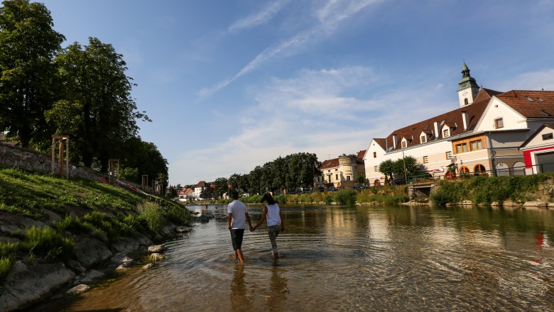 Erlaufhafen, © schwarz-koenig.at A couple walks hand in hand through a shallow river in a town with historic buildings and trees.