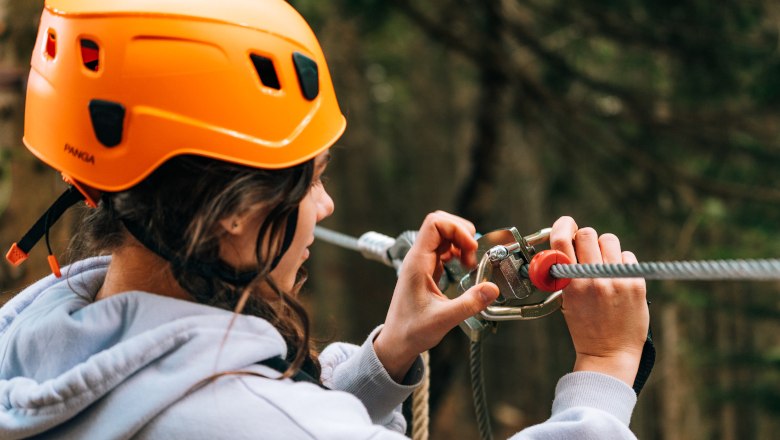 Hirschenkogel forest ropes course Semmering, © Semmering-Hirschenkogel Person wearing an orange helmet attaches safety equipment to a rope in the forest ropes course.
