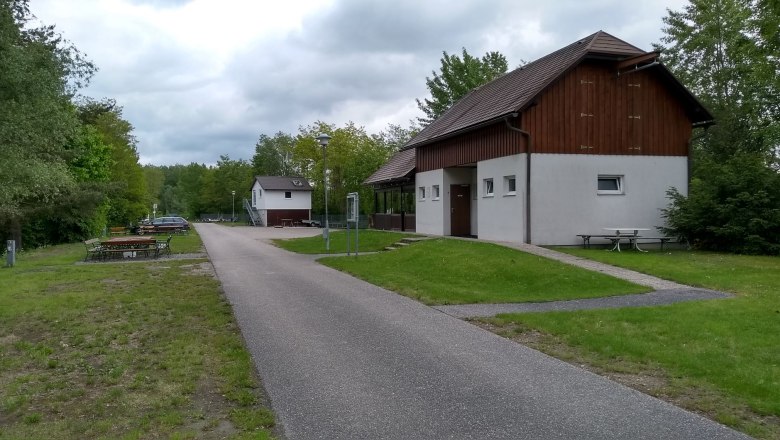 Boathouse in Emmersdorf, © Amt der NÖ Landesregierung, Abt. Wasserwirtschaft /Ing. Werner Hauer A path leads past a white building with wooden cladding, surrounded by trees and meadows, under a cloudy sky.
