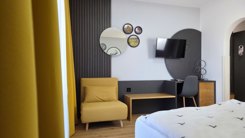 Modern "feel-good room", © Julian Weber, Café Pension Kristall Modernly furnished room with a yellow armchair, mirrors on the wall, a TV and a desk with chair.