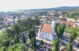 Kunigundenkirche with surrounding cemetery, © Marktgemeinde Mailberg Aerial view of a church with a red roof, surrounded by a cemetery and trees, in a rural setting with houses and hills in the background.