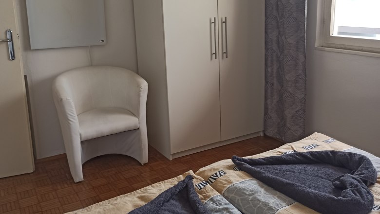 Guest room, © Multimedia Lauda A guest room with two beds, a white wardrobe, an armchair and a window with a curtain.