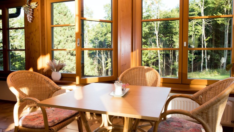 Table in the breakfast room, © Dorfschmiede eG A table with four rattan chairs in a sunny room with large windows and a view of a forest.