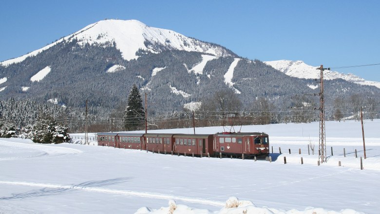 Ötscherbär family adventure train, © PROKOP A train travels through a snowy landscape with a snow-covered mountain in the background.