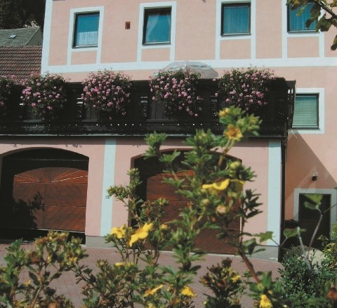 Guesthouse Thaya, © Gästehaus Thaya A two-storey pink building with flowering plants on the balcony and yellow flowers in the foreground.