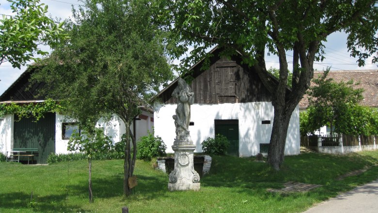 Wine cellar lane Großkadolz, © Gemeinde Seefeld-Kadolz A white building with a wooden gate and statue in the foreground, surrounded by trees and green lawns.