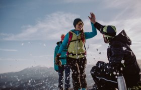 Unterberg ski area, © Schigebiet Unterberg | katischweiger.fotography Three people in the snow, one high-fiving another, with mountains in the background.