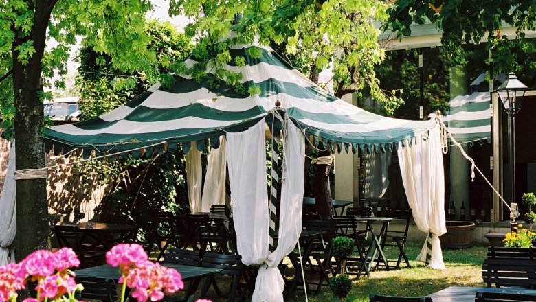 Tent in the garden, © Weingut Fischer A striped tent with white curtains stands in a garden surrounded by trees and flowers.