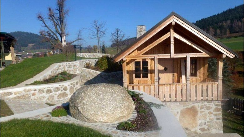 EXPERIENCE: Stone & Garden, © Familie Anderl Small wooden house in a garden with stone walls and a large rock.