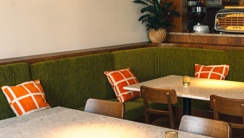 Table inside restaurant, © Womo Interior view of a stylish café with a green bench, orange and white cushions, wooden furniture and an illuminated wall lamp.